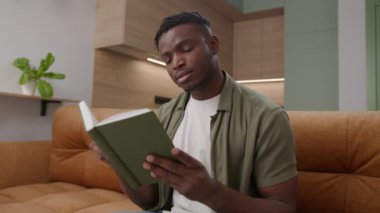 Siyah Adam Evdeki Koltukta Oturarak Kitap Okuyor. Afrika kökenli Amerikalı erkek oturma odasında kağıt kitap okuyor. 4K