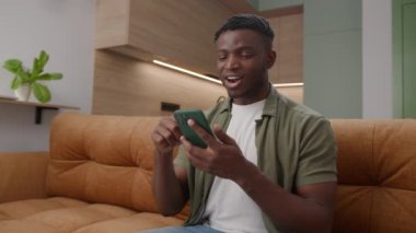 Siyah adam cep telefonundan iyi haberler alıyor. Evdeki koltukta oturan akıllı telefonla gülümseyen Afro-Amerikalı bir adam. 4K