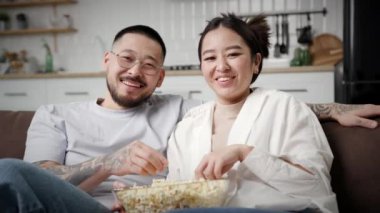 TV izlerken gülen ve patlamış mısır yiyen Asyalı çift. Aile kanepede otururken eğleniyor. Kanepede kameraya bakarak mutlu bir eş ve koca. 4K