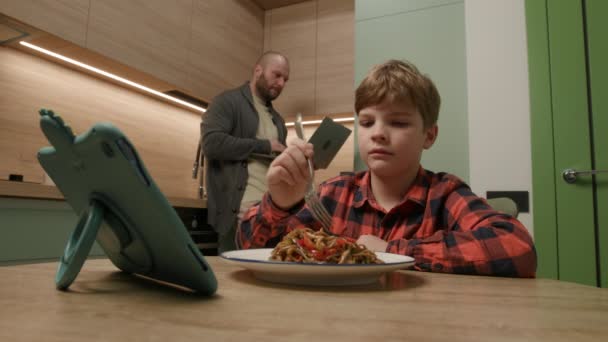 男の子はタブレットで食事を楽しんでいますが パパはラップトップでマルチタスクします テクノロジーと現代の家族生活のダイナミクスを説明するのに最適 — ストック動画