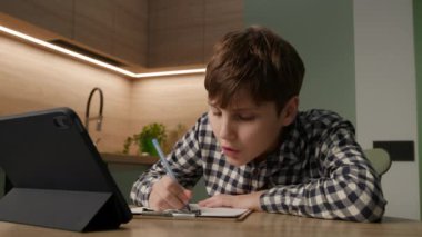 Çalışkan öğrenci bir kalemle yazıyor, online ödevlerine dalmış, tabletine bakıyor. Modern çalışma yöntemleri ve teknolojik entegre öğrenme