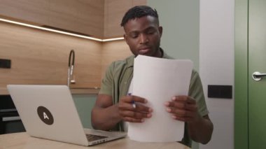 Görevleri iyi bir şekilde yöneten siyah bir adam, kağıt belgeleriyle birlikte dizüstü bilgisayarını kullanarak, işini evden sorunsuz bir şekilde yürütüyor. 