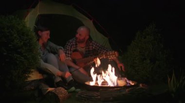 Yıldızlı gökyüzünün altında, bir adam kamp ateşinin yanında eşine serenat yapıyor, kamp kaçamaklarında romantizmi ateşliyor.