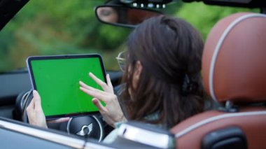 Çoklu görevlerde bulunan bir kadın, arabasında otururken yeşil ekran tableti kullanır ve bu da mobil verimliliği gösterir..