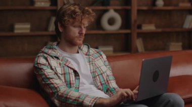 Kıvırcık saçlı, sıradan iş kıyafetleri içinde odaklanmış bir adam, rahat bir ofis ortamında laptopuyla dikkatle çalışıyor..