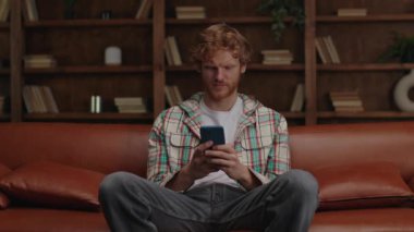Rahat genç bir adam, kızıl saçlı, şık bir iç mekanda deri koltukta otururken telefonunu karıştırıyor..
