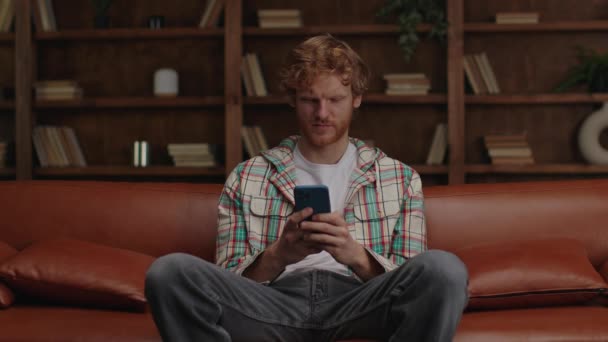 Розслаблений Молодий Чоловік Імбирним Волоссям Переглядає Свій Телефон Сидячи Шкіряному — стокове відео