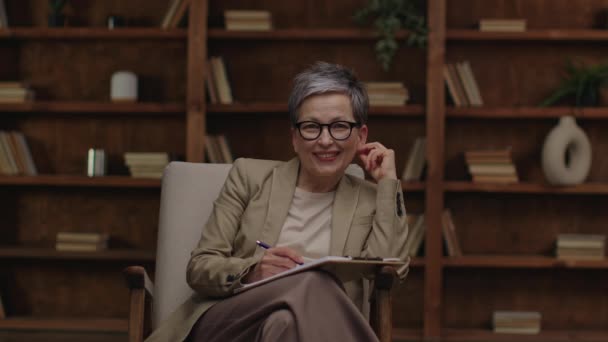 Munter Kvinnelig Terapeut Med Briller Sitter Stol Noterer Med Bibliotek – stockvideo