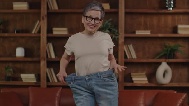 Lächelnde Seniorin Prahlt Mit Gewichtsverlust Indem Sie Große Jeans Hochhält — Stockvideo