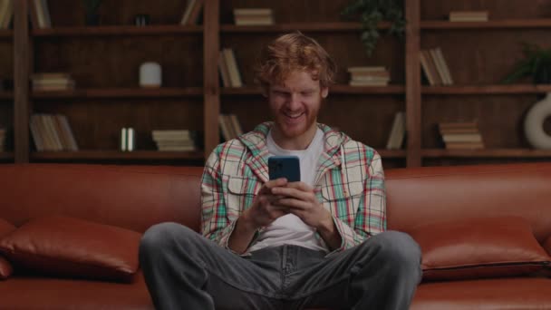 Młody Człowiek Śmiejący Się Patrząc Smartfona Siedzący Skórzanej Kanapie — Wideo stockowe