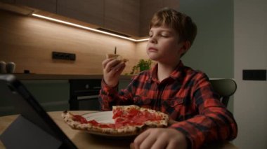 Çocuk bir tablet bilgisayarın içindeyken lezzetli bir pizza diliminden zevk alıyor. Teknoloji ve yemek zamanının modern karışımını tasvir etmek için mükemmel.