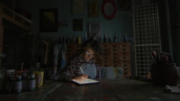 在一个灯光昏暗的工作室里 艺术家在平板电脑上素描 四周都是艺术品 — 图库视频影像