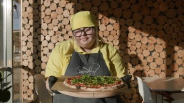 Down Sendromlu bir kafe çalışanı pizzayı sıcak bir şekilde sunar. Eklemeyi ve mutfak neşesini kutlar. Çeşitliliği kucaklama.