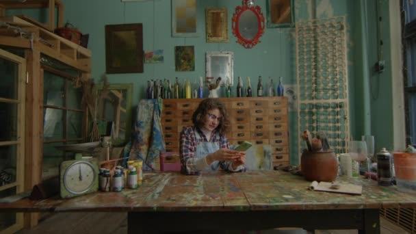 Curly Haired Artist Browsing Smartphone Amidst Vibrant Art Studio Setting — Stock Video