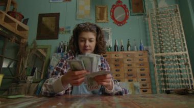 Stüdyoda resim malzemeleriyle çevrili kıvırcık saçlı bir sanatçı para sayıyor.