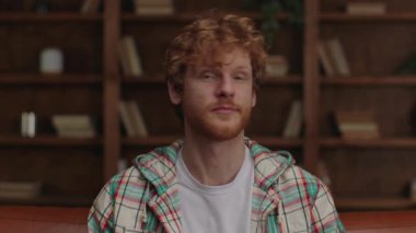 Kıvırcık kızıl saçlı neşeli bir adam sıcacık bir kütüphanedeki kahverengi deri koltukta rahatça oturuyor ve gülümsüyor..