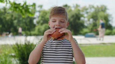Canlı bir şehir parkında, 8 yaşındaki bir çocuk lezzetli bir burgerin tadını çıkarıyor, hareketli bir şehir ortamında açık havada yemek yemenin zevkini yaşıyor.