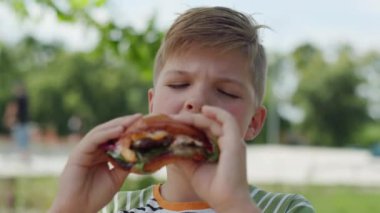 Canlı bir şehir parkında, 8 yaşındaki bir çocuk lezzetli bir burgerin tadını çıkarıyor, hareketli bir şehir ortamında açık havada yemek yemenin zevkini yaşıyor.