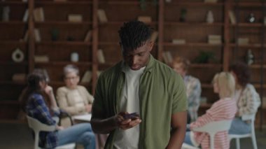 Arka planda modern teknoloji ve destek ortamlarının karışımını vurgulayan bir grup terapi seansı gerçekleşirken, siyahi bir adam telefonuna kayıyor.