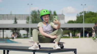 Kask takan bir çocuk şehir parkında kaykayının üzerinde oturuyor, cep telefonunu video görüşmesi için kullanıyor, modern teknolojiyle açık hava oyunlarını birleştiriyor.