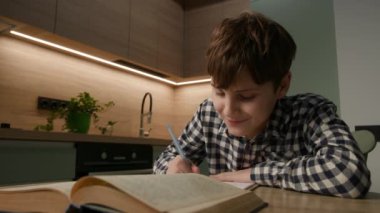 Genç bir çocuk evde oturuyor, bir kitaba gönderme yaparken bir deftere yazıyor, odaklanmış bir çalışma anını yakalıyor ve ev ortamında öğreniyor..