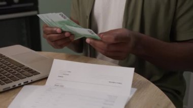 Black 'in yakın plan fotoğrafları bir fatura üzerinden Avro banknotlarını sayarken aynı zamanda bir dizüstü bilgisayarda yazarak, finansal yönetimi ve modern bir ortamda birden fazla görevi ön plana çıkarıyor