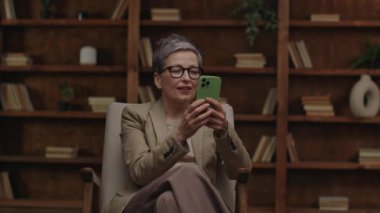 Gözlüklü neşeli olgun bir kadın, telefonuna bakarken kazanan bir jest yapıyor..