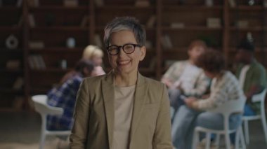 Gülümseyen kıdemli bir terapist kollarını kavuşturmuş bir şekilde ayakta dururken, arka planda beş kişilik bir destek grubu liderlik ve grup terapisi anlarını yakalıyor.