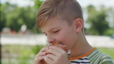 Canlı bir şehir parkında, 8 yaşındaki bir çocuk lezzetli bir burgerin tadını çıkarıyor, hareketli bir şehir ortamında açık havada yemek yemenin zevkini yaşıyor.