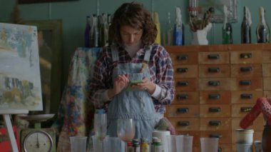 Yaratıcı bir kadın atölyede duruyor, akıllı telefonuyla interneti karıştırıyor. Aletler ve ilhamlarla dolu bir çalışma alanında samimi bir odak ve teknoloji anı.