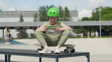 Kask takan bir çocuk, şehir parkında kaykay yaparken cep telefonuyla maç kazanmasını kutluyor. Açık hava oyunlarında bir neşe ve başarı anı yakalıyor.