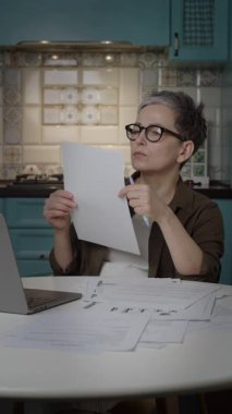 Olgun bir kadın evde dizüstü bilgisayar kullanırken evrak işlerini halleder. Uzaktan çalışma, online bankacılık, muhasebe, finansal planlama ve belge yönetimi konuları için mükemmel. Dikey ekran 