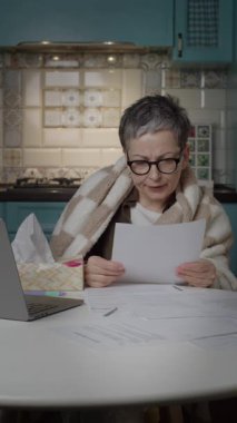 Hasta, olgun bir kadın evde çalışıyor. Laptop kullanırken öksürüyor ve battaniyeye sarılıyor. Uzaktan çalışma, hastalık, grip ve iş hayatı dengesi temaları için mükemmel