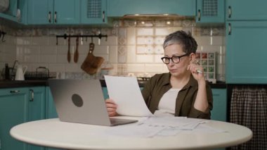 Olgun bir kadın evde dizüstü bilgisayar kullanırken evrak işlerini halleder. Uzak çalışma, çevrimiçi bankacılık, muhasebe, finansal planlama ve belge yönetimi konuları için mükemmel