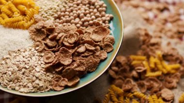 Ekmek, pirinç, kepekli taneler, yulaf, buğday ve mısır makarnası da dahil olmak üzere çeşitli karbonhidrat zengini yiyecekler bir tabakta sergilenmekte ve ahşap bir masada dönmektedir. Beslenme ve sağlıklı beslenme için mükemmel.