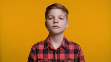 Genç bir çocuğun yüzünde ciddi bir ifade var, hafif kaşları çatık ve doğrudan kameraya bakıyor. Arkaplan koyu sarı renktedir..