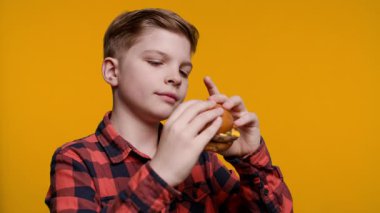 Genç bir çocuk yüzünde kocaman bir gülümsemeyle lezzetli bir burgerin tadını çıkarıyor. Parlak sarı arka plan onun yemek saatine eğlenceli ve canlı bir dokunuş katıyor..