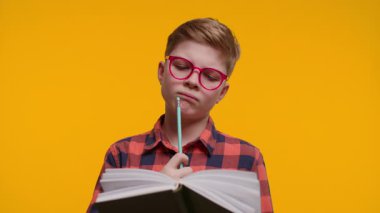 Gözlük takan genç bir çocuk elinde kalem tutarken derin bir konsantrasyonla kitap okur, not almaya hazırdır. Parlak sarı arka plan, onun öğrenmeye olan ilgisini vurguluyor..