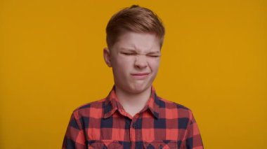Genç bir çocuk, tatminsizlik ve nezaketin bir karışımını göstererek kameraya beceriksizce gülümsüyor. Onun ifadesi parlak ve neşeli sarı arka planla çelişiyor..