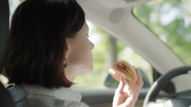 Bir kadın hamburger yerken arabasını sürüyor, direksiyonun başındaki bir anlık dikkat dağınıklığını gösteriyor ve araba kullanırken birden fazla iş yapmanın risklerini vurguluyor..