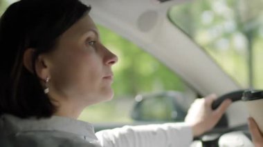 Bir kadın arabasını sürerken kahvesini yudumlar, tipik bir sabah işe gidiş gelişi anını yakalar ve sürücünün birden fazla işi vardır..