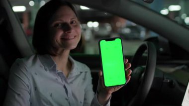 Park halindeki arabasında oturan bir kadın, elinde yeşil ekran akıllı bir telefon ile, kişiselleştirilebilir içerik veya uygulama modellemesi için hazır bekliyor..