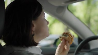 Bir kadın hamburger yerken arabasını sürüyor, direksiyonun başındaki bir anlık dikkat dağınıklığını gösteriyor ve araba kullanırken birden fazla iş yapmanın risklerini vurguluyor..