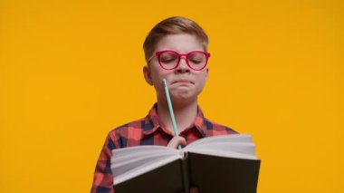 Gözlük takan genç bir çocuk elinde kalem tutarken derin bir konsantrasyonla kitap okur, not almaya hazırdır. Parlak sarı arka plan, onun öğrenmeye olan ilgisini vurguluyor..