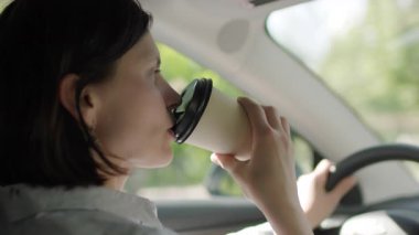 Bir kadın arabasını sürerken kahvesini yudumlar, tipik bir sabah işe gidiş gelişi anını yakalar ve sürücünün birden fazla işi vardır..