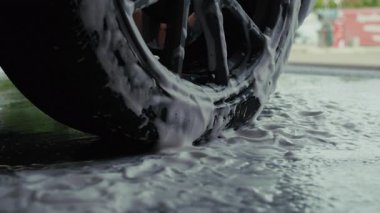 Açık hava araba yıkama sırasında kalın sabunlu köpükle kaplanmış bir araba tekerleğinin yakın plan çekimi temizlik sürecini ve köpüklü köpükleri vurguluyor..