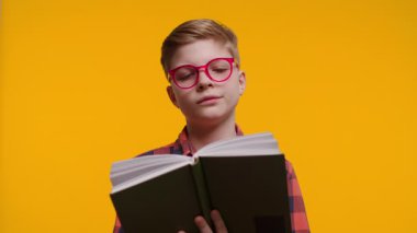 Gözlüklü neşeli bir çocuk neşe ve ilgi dolu bir kitap okur. Parlak gülüşü ve odaklanmış ifadesi parlak sarı arka plana çarpıyor..