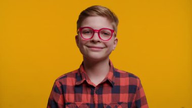 Gözlüklü genç bir çocuk kameraya sıcak bir şekilde gülümsüyor, mutluluk ve güven saçıyor. Parlak sarı arka plan, onun neşeli ifadesini ve neşeli bakışını vurguluyor..