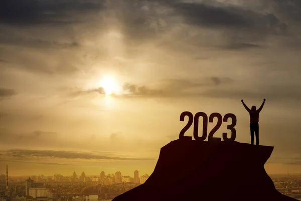 The concept of Victory in the new year 2023. A man stands with his hands raised up against the background of a sunset the number 2023.