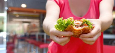 Kadının biri, kafenin bulanık arka planında hamburger gösteriyor..